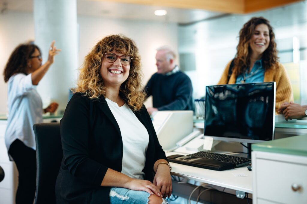 Portrait of a smiling woman sitting at her desk with people in background.